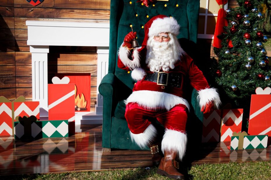 Santa Claus sitting cheerfully amidst festive holiday decorations, ringing a bell.